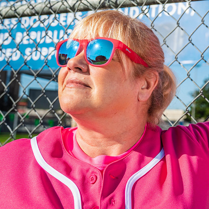 Una donna con un'uniforme da baseball rosa caldo e occhiali da sole larghi rosa caldo con lenti blu si appoggia a una recinzione in un raggio di sole.