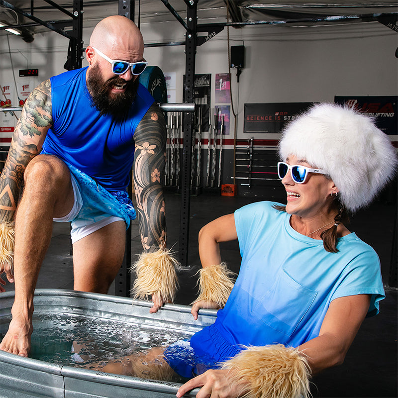 Un uomo tatuato si immerge in un bagno di ghiaccio accanto a una donna con un cappello di pelliccia bianco, entrambi con occhiali da sole bianchi con lenti blu.