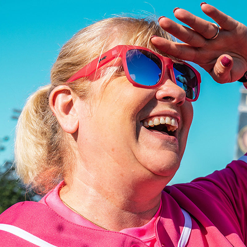 Vista di tre quarti di una donna in uniforme da baseball rosa e occhiali da sole rosa con lenti blu che sorride su un campo da baseball.
