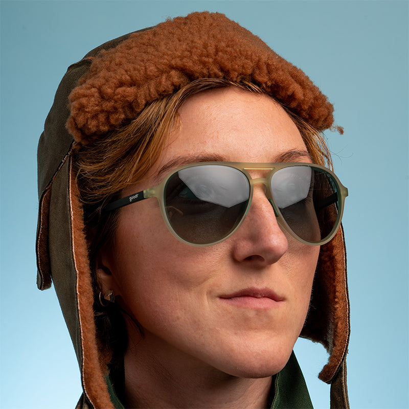 Una donna con un berretto da aviatore marrone sfocato guarda con sicurezza, indossando aviatori verde cadetto con lenti verdi sfumate.