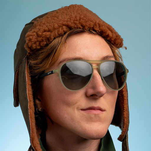 Una donna con un berretto da aviatore marrone sfocato guarda con sicurezza, indossando aviatori verde cadetto con lenti verdi sfumate.