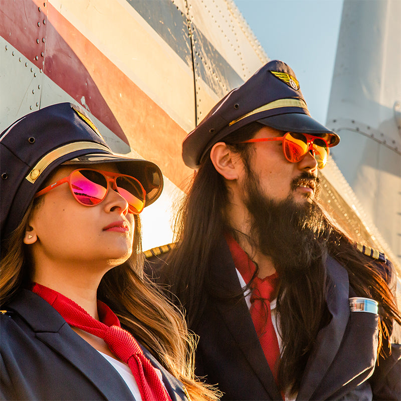 Due piloti spettinati che indossano occhiali da aviatore con lenti rosse si appoggiano a un aereo e guardano il tramonto.