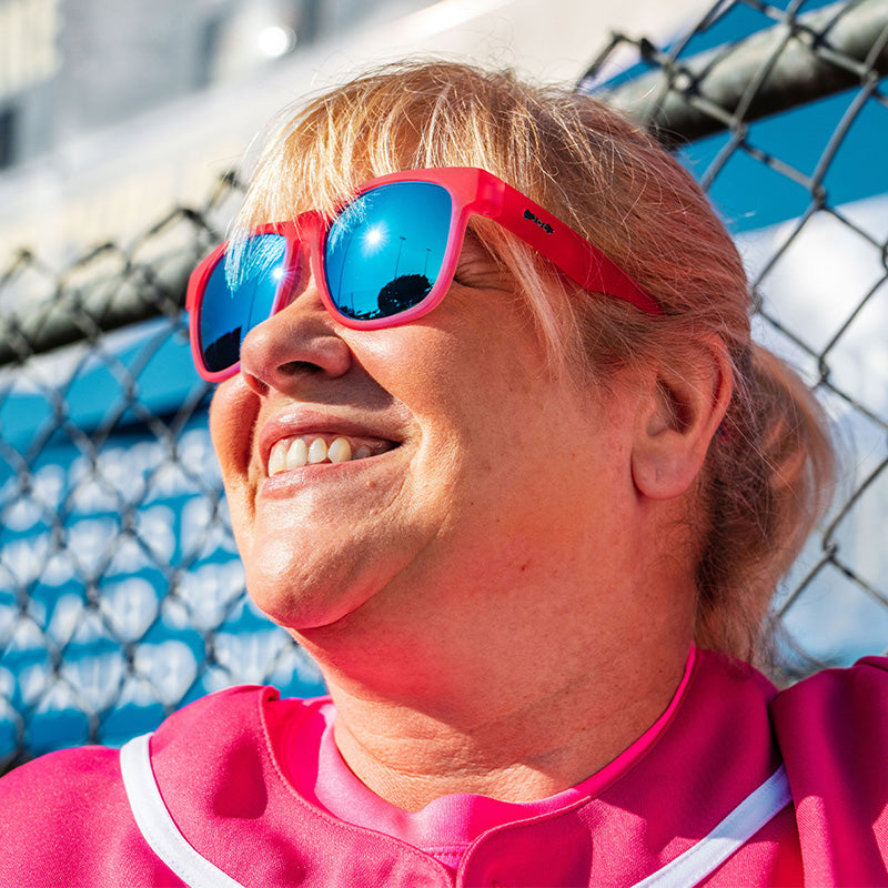 Una donna in uniforme da baseball rosa e occhiali da sole rosa con lenti blu riflettenti sorride, appoggiata a una recinzione.