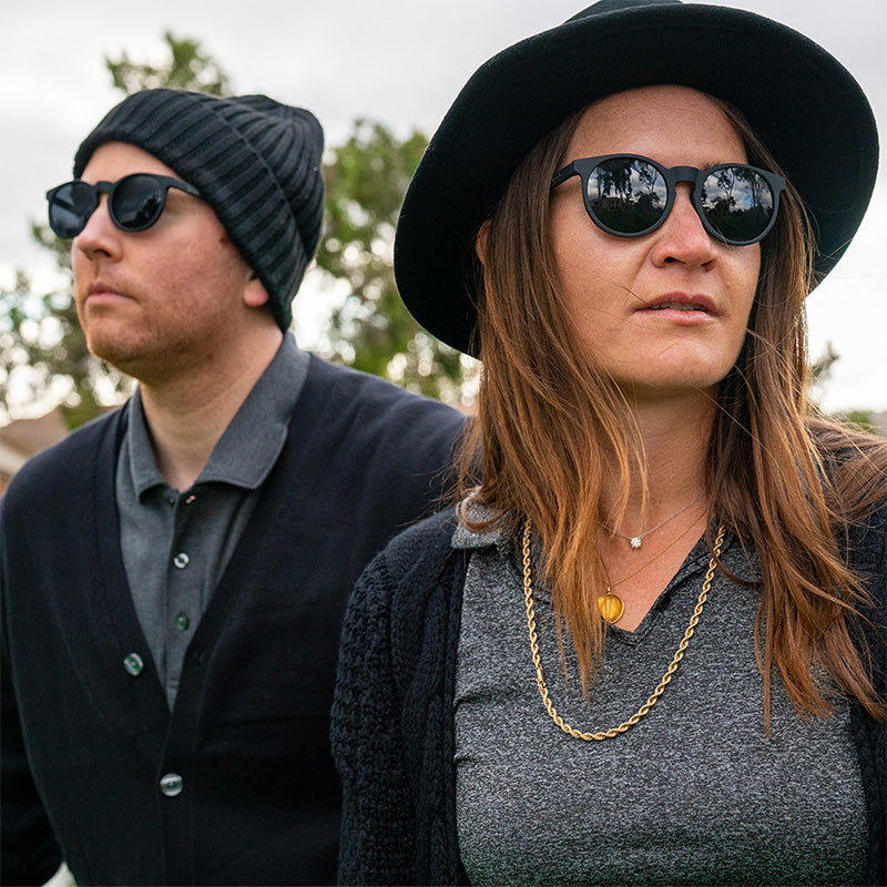 Un uomo e una donna hipster con abiti neri chic indossano occhiali da sole neri rotondi con lenti nere non riflettenti.