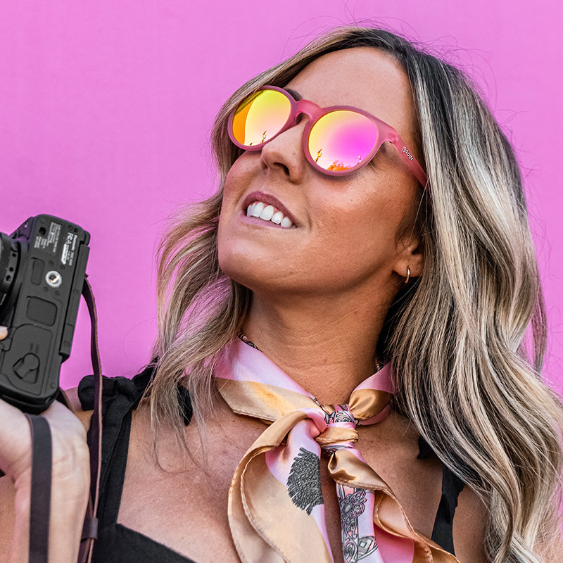 Una donna elegante che indossa occhiali da sole rotondi rosa e tiene in mano una macchina fotografica guarda di lato, con un muro rosa caldo alle spalle.