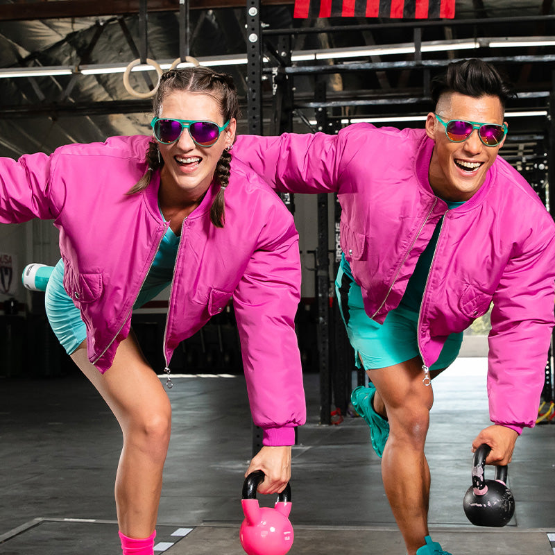 Un uomo e una donna sorridenti in bomber rosa e aviatori verde acqua con lenti riflettenti rosa si allenano con i kettlebell.