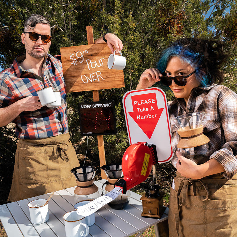 Due baristi hipster che indossano occhiali da sole rotondi con lenti marroni posano in un chiosco di caffè pour-over indulgentemente artigianale.