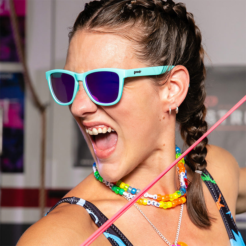 Una donna con collane di plastica arcobaleno e occhiali da sole blu baby con lenti riflettenti viola grugnisce ferocemente in una palestra.