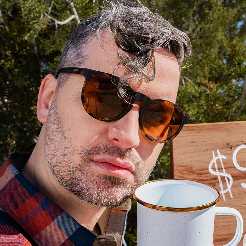 Un barista hipster ci guarda con giudizio, indossando occhiali da sole marroni tartarugati con lenti marroni non riflettenti.