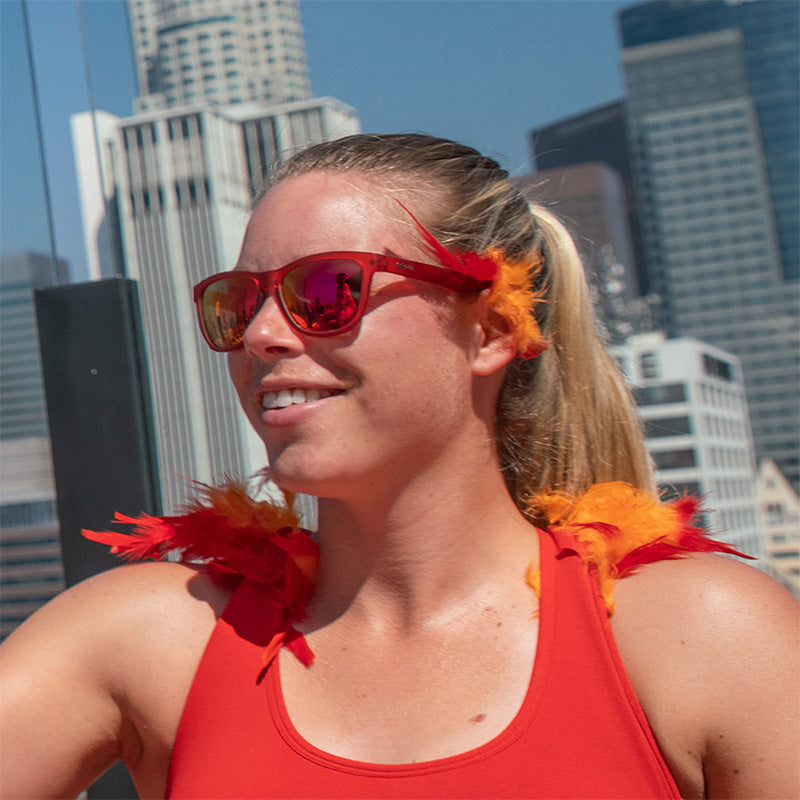 Una donna che indossa occhiali da sole rossi con lenti rosse e accenti di piume rosse e arancioni, con alle spalle lo skyline della città.
