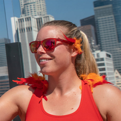 Una donna che indossa occhiali da sole rossi con lenti rosse e accenti di piume rosse e arancioni, con alle spalle lo skyline della città.
